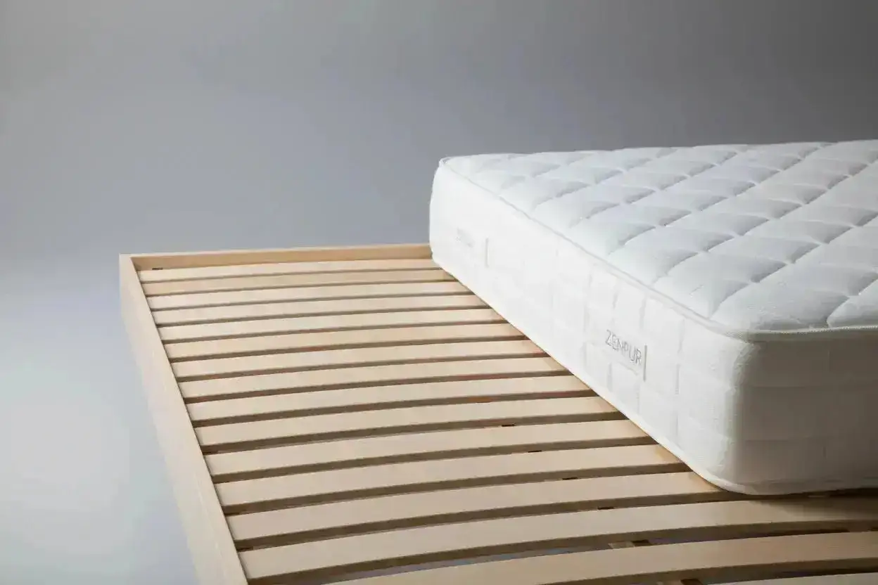 Un matelas trop grand pour sommier, ses lattes de bois en premier plan, puis le matelas blanc matelassé.