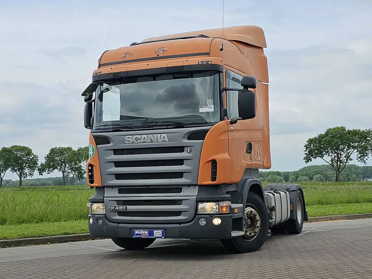 Pomarańczowa Scania R420 z szarym grillem stoi na tle zielonych pól i drzew.