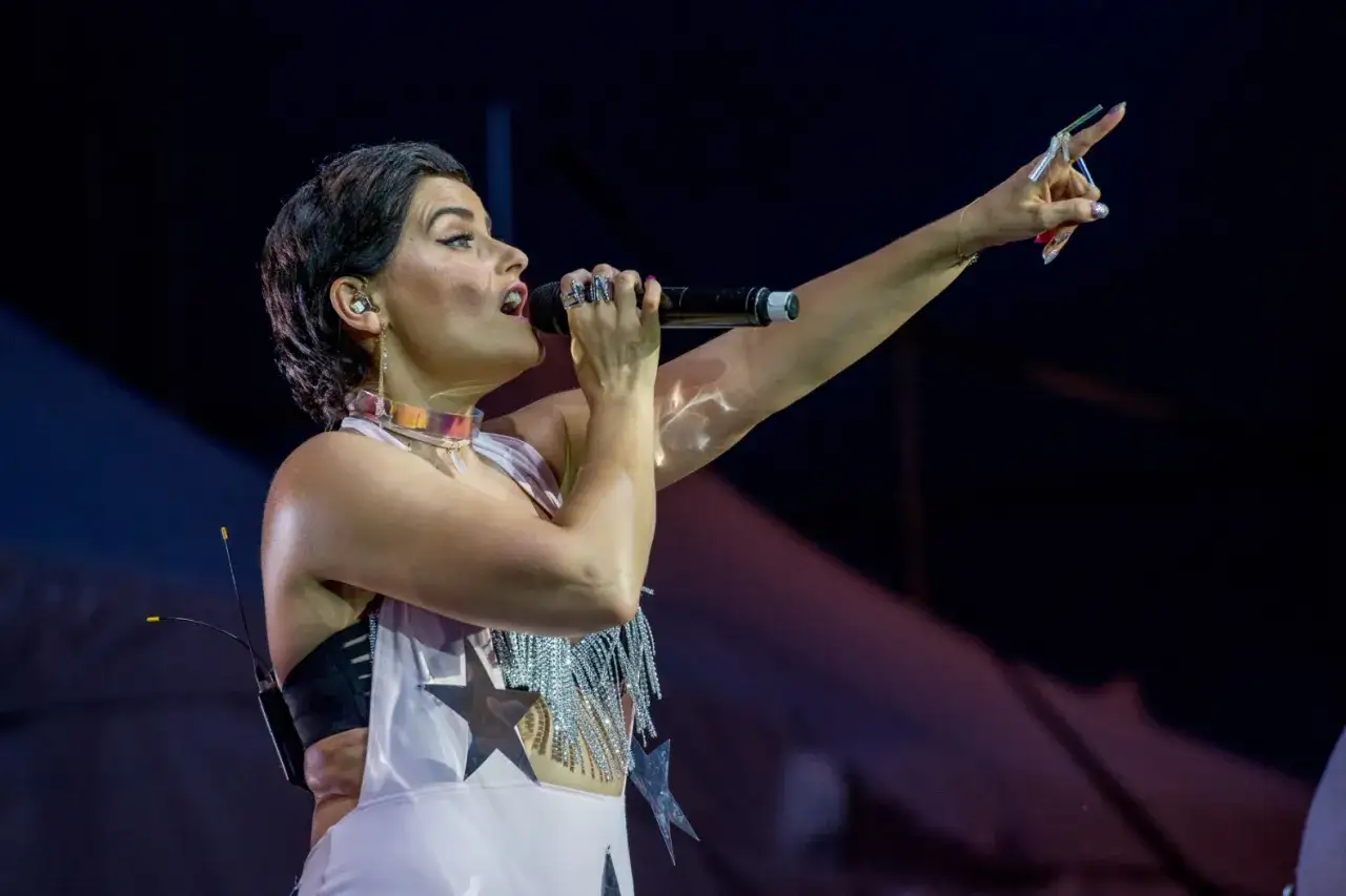 Nelly Furtado 2024 na scenie, śpiewa do mikrofonu, z podniesionym palcem wskazującym, w błyszczącym stroju z gwiazdami.