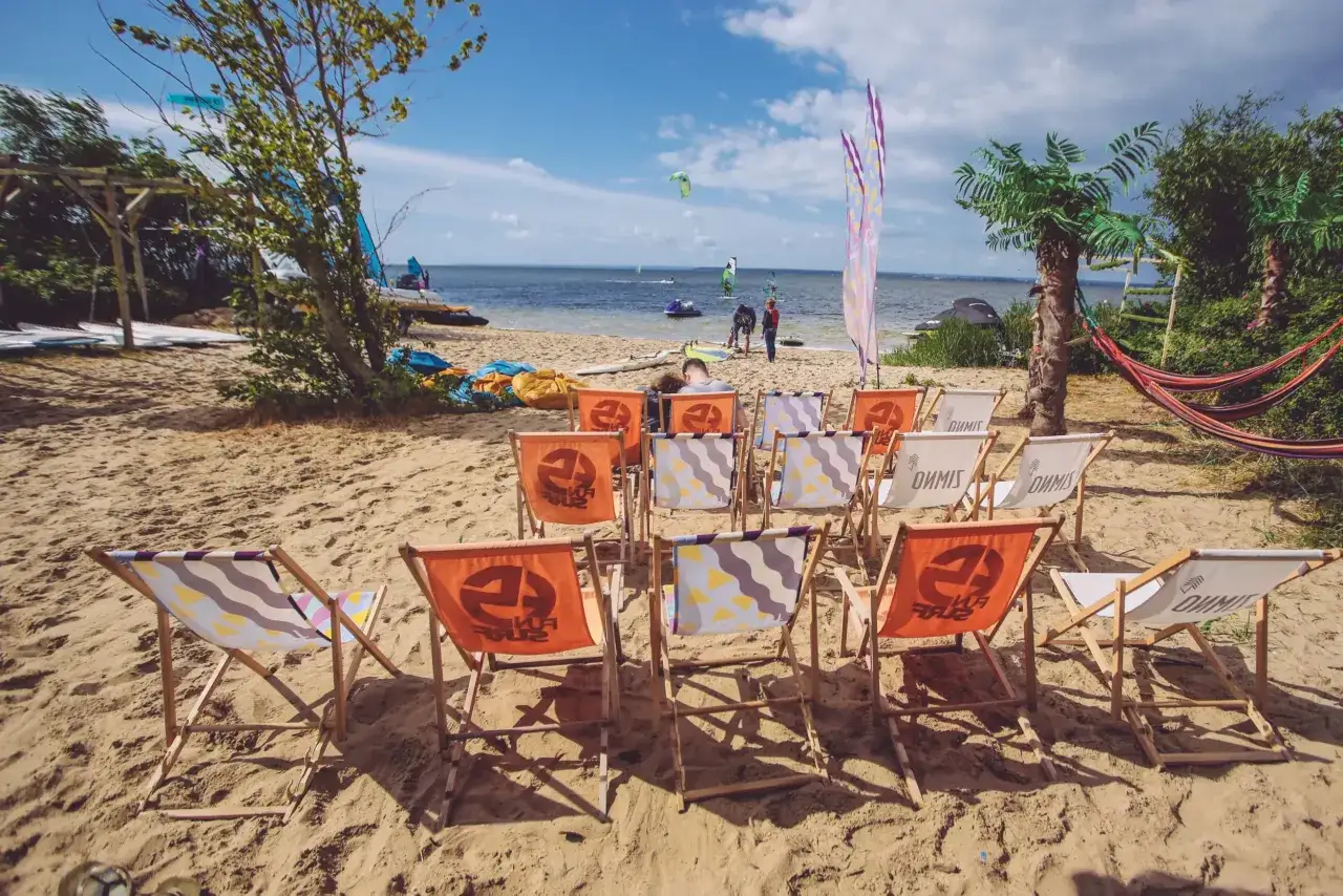 Kemping Polaris Chałupy zaprasza na plażę z leżakami i widokiem na wodę, gdzie można podziwiać windsurferów.