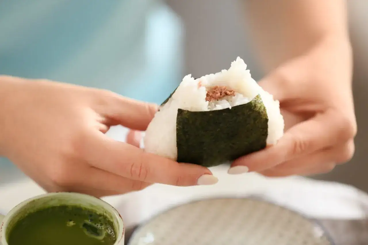 Hands hold a tuna-filled onigiri, showing its perfect size for a snack. A cup of matcha tea sits nearby.