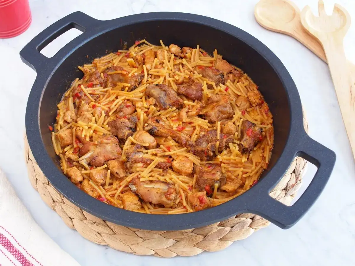 Un plato humeante de fideos a la cazuela con trozos de pollo y costillas de cerdo, servido en una paellera negra sobre una esterilla de mimbre.