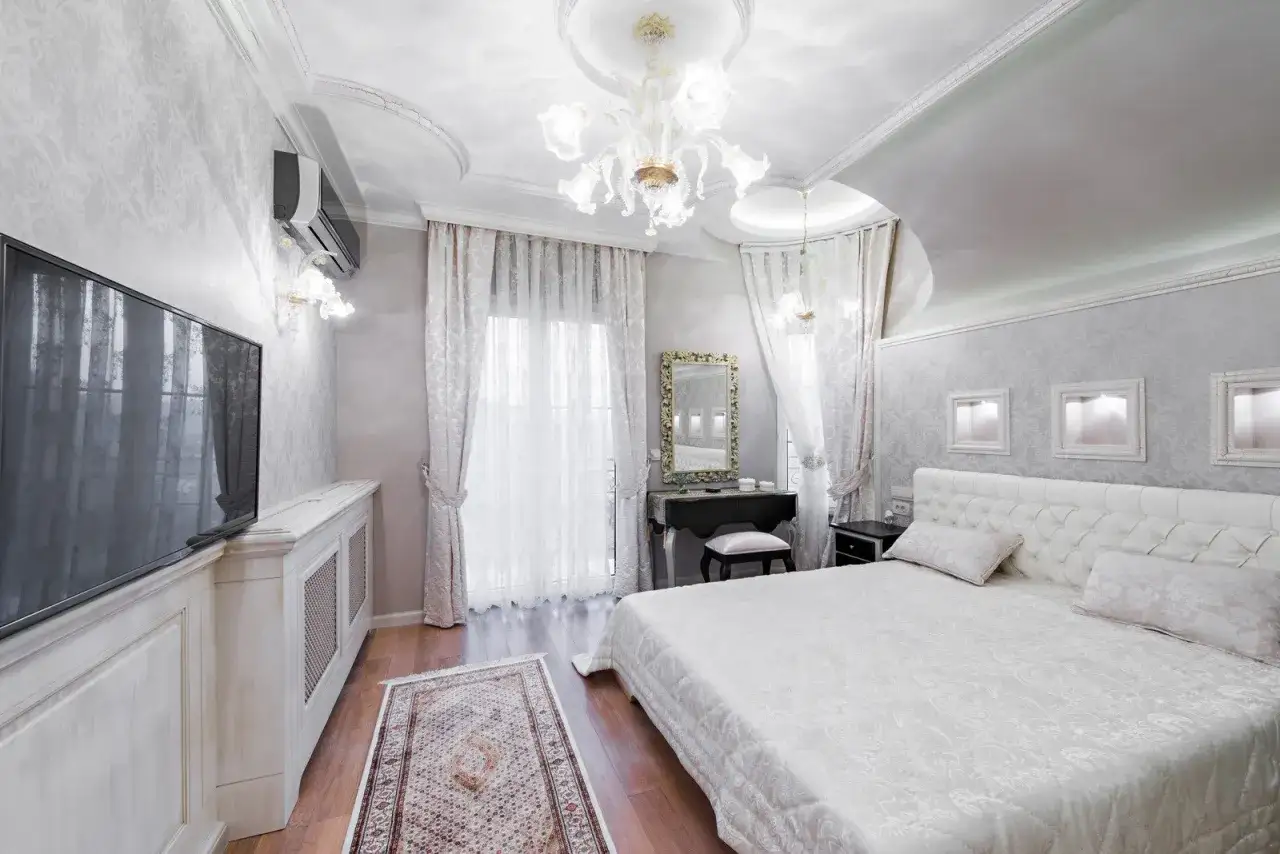 Elegancka, mała sypialnia w stylu glamour z białym łóżkiem, toaletką i ozdobnym żyrandolem.