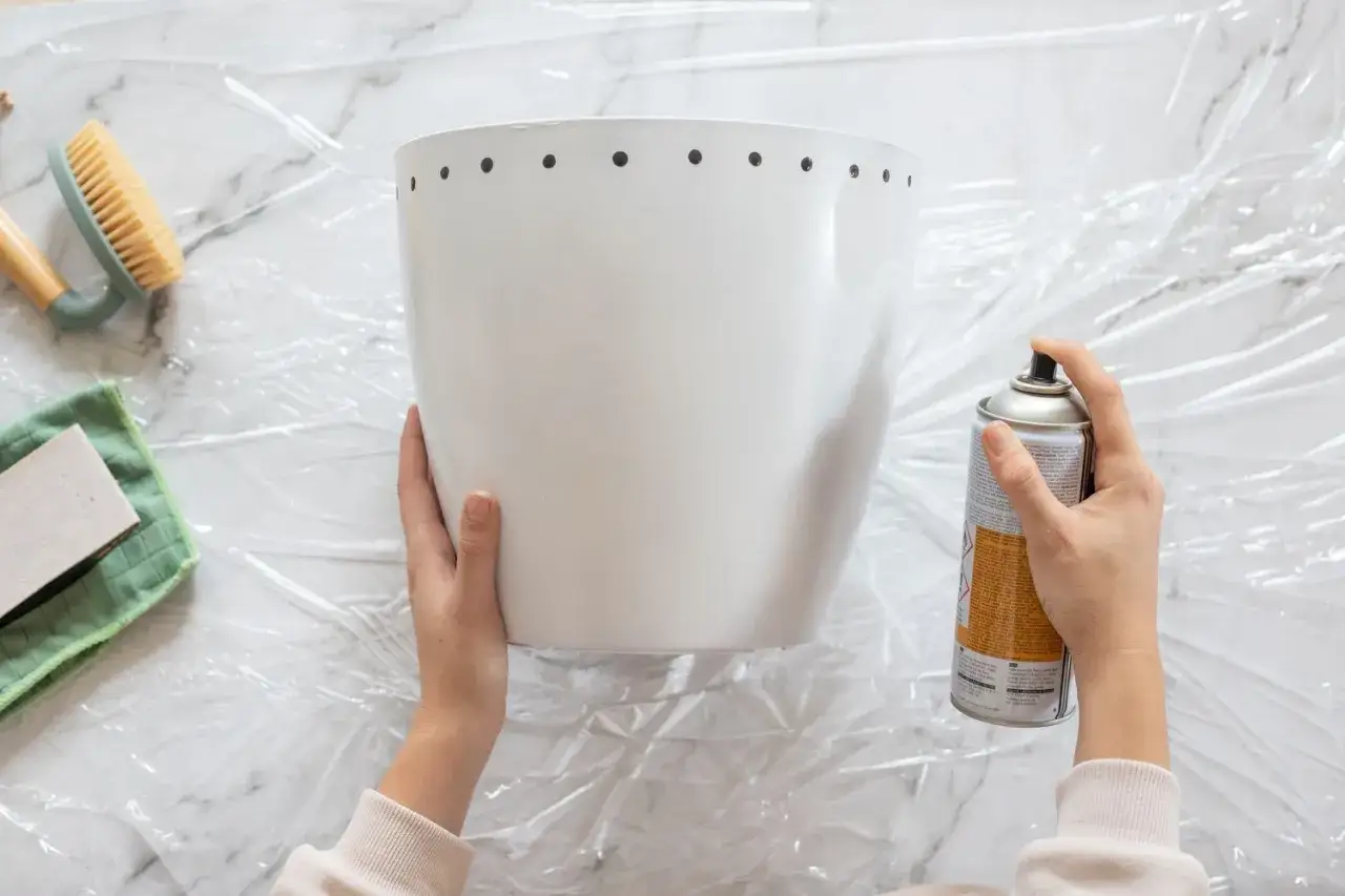 Manos rociando pintura para pintar plástico en una maceta blanca. A su lado, un cepillo y una lija.