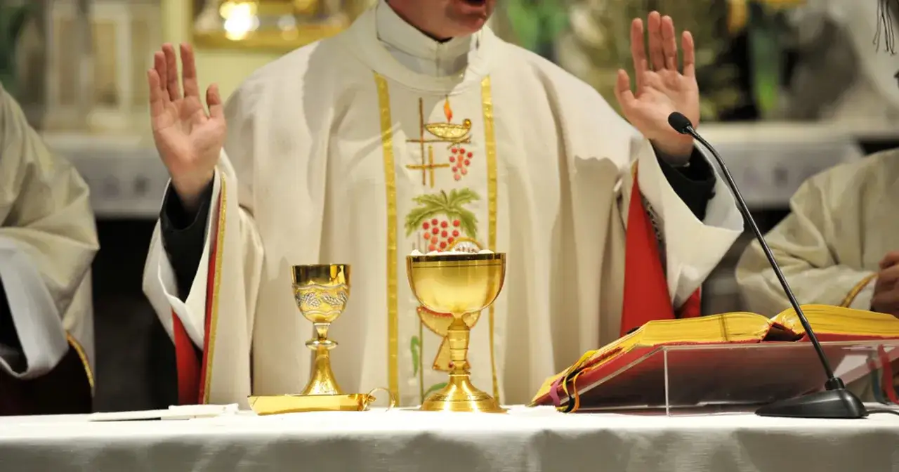 Kapłan w białej szacie z haftem winogron i kielichami podczas mszy. W tle widać mikrofon i księgę, co sugeruje uroczystość i dyspensa.