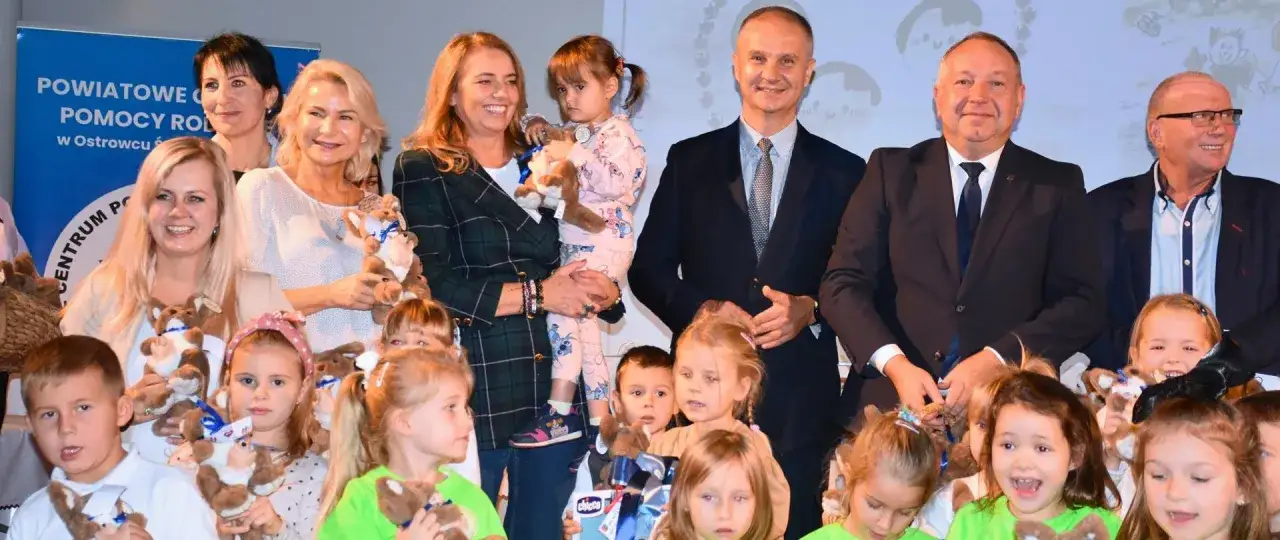 Dzieci z pluszakami, wśród nich mała córka po francusku, w otoczeniu dorosłych.