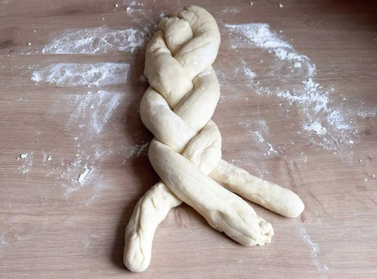 Ein frisch geflochtener Zopf Brot liegt auf einem Holztisch, best&auml;ubt mit Mehl.