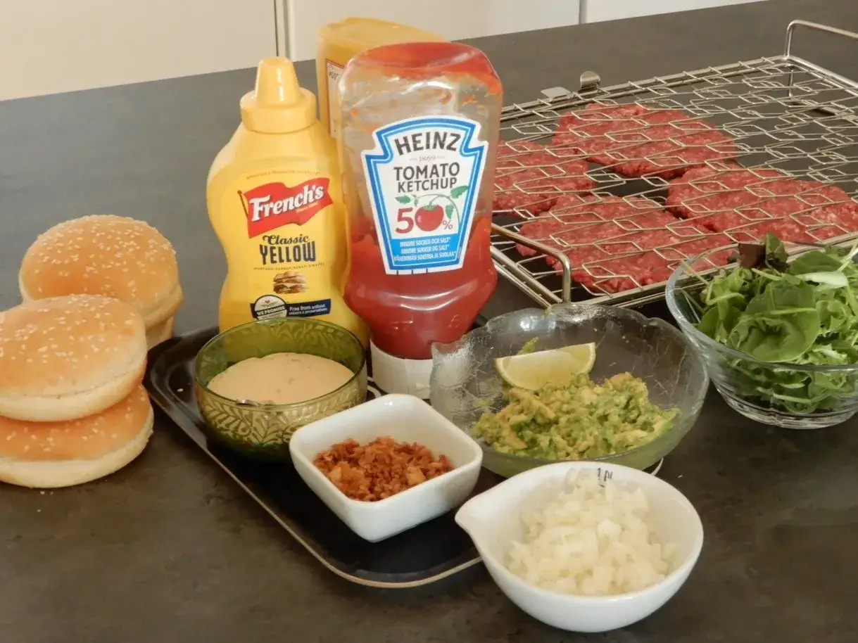 F&ouml;rberedelser f&ouml;r att grilla hamburgare: biffar p&aring; grillgaller, hamburgerbr&ouml;d, ketchup, senap, guacamole, l&ouml;k, sallad och dressing.