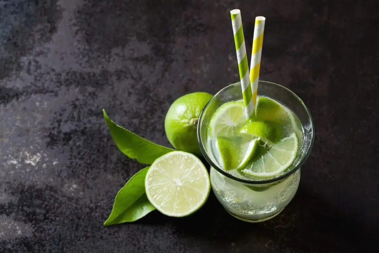 Orzeźwiający drink z limonką, udekorowany plasterkami owocu i kolorowymi słomkami. Obok leżą świeże limonki i liście.