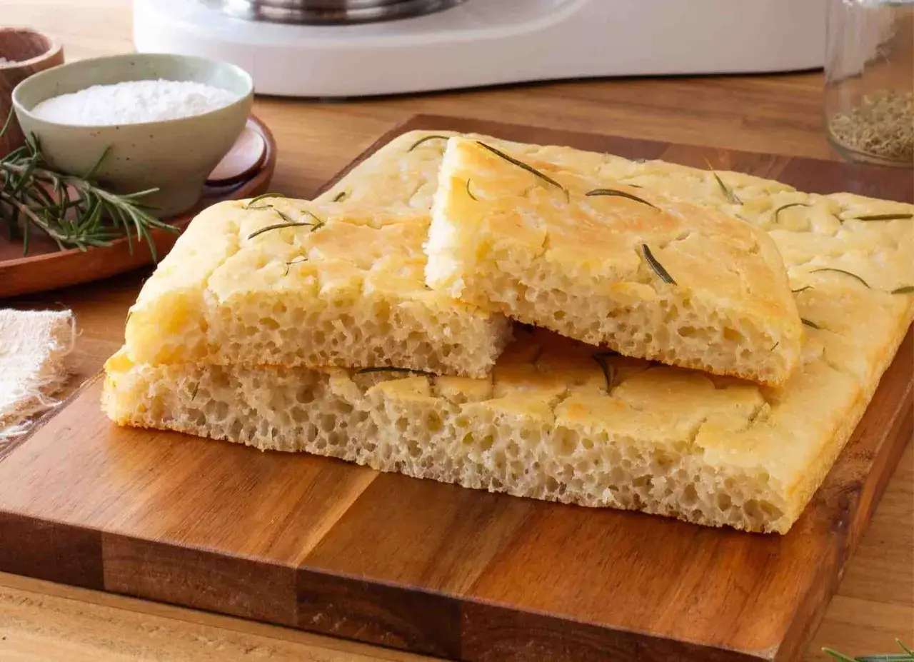Focaccia senza glutine soffice e dorata, guarnita con rosmarino fresco, su un tagliere di legno.