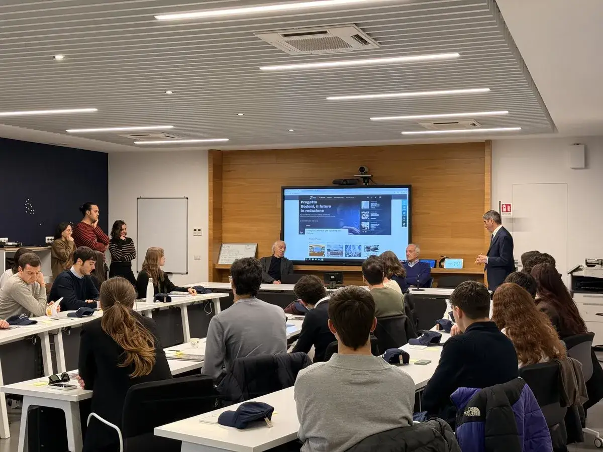 Aula di una scuola di giornalismo con studenti e docenti che ascoltano una presentazione su un grande schermo.