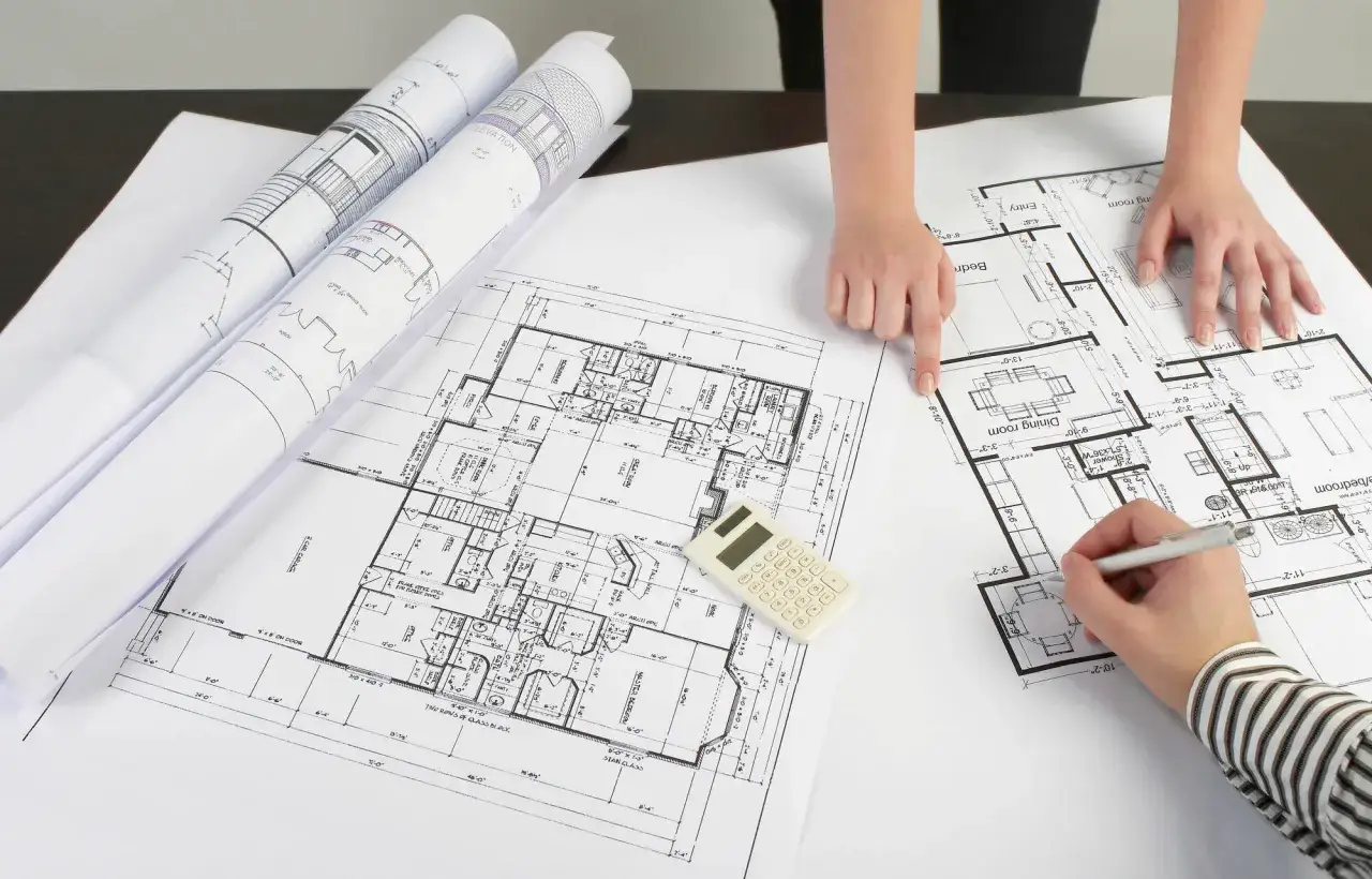 Rysunki techniczne i plany domu na stole. Dwie osoby analizują adaptację projektu budowlanego, jedna wskazuje palcem, druga pisze.