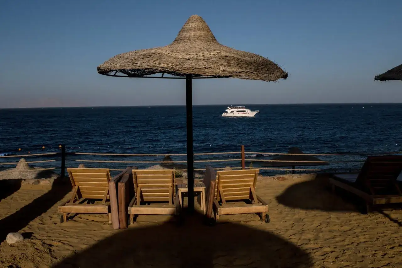 Plaża w Szarm el-Szejk z leżakami i parasolem. Na morzu widać białą łódź. Czy w Szarm el-Szejk jest bezpiecznie? Tak, wygląda na spokojne miejsce na wypoczynek.