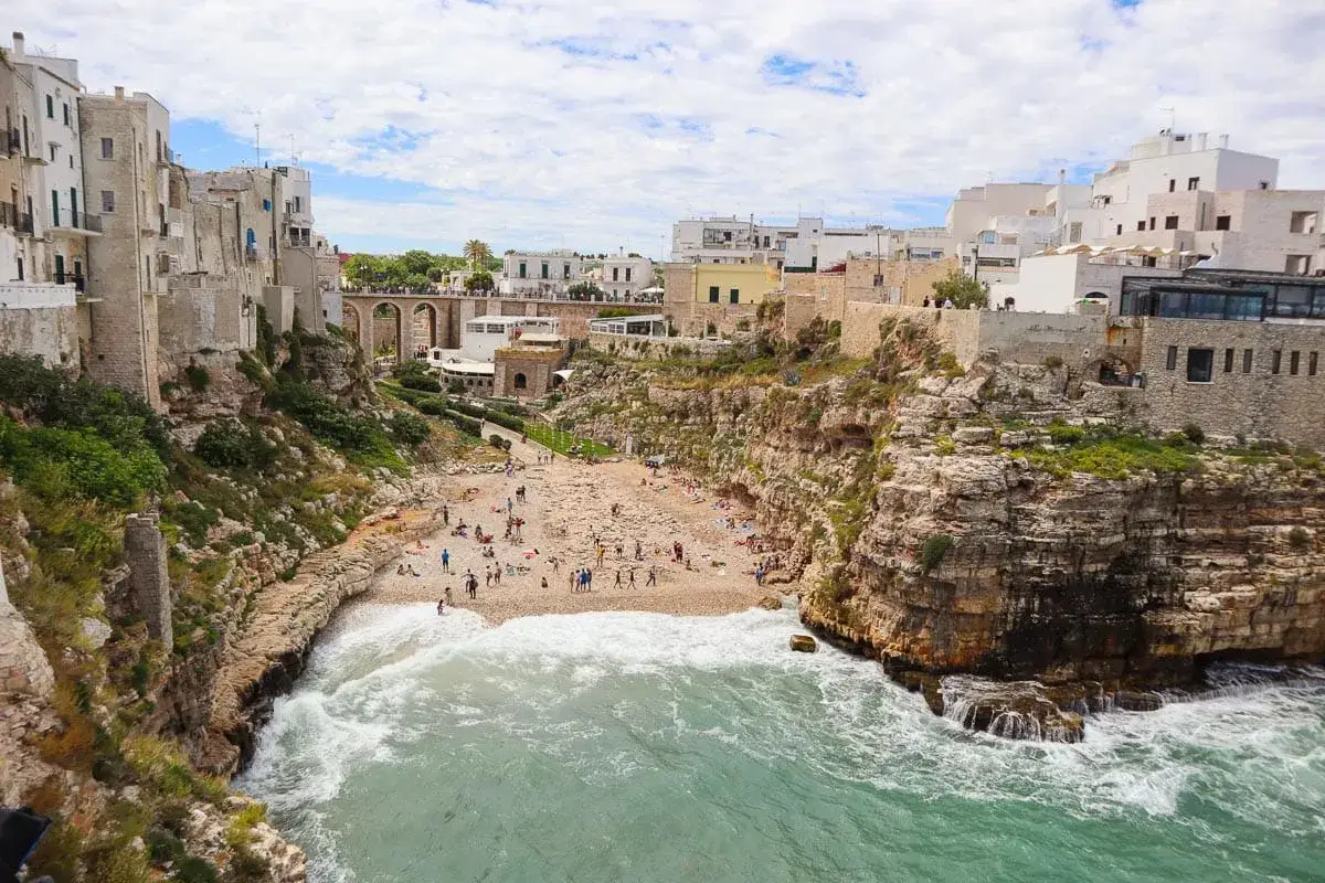 A picturesque beach nestled between cliffs, perfect for a southern Italy trip itinerary. White buildings perch above, overlooking sunbathers.