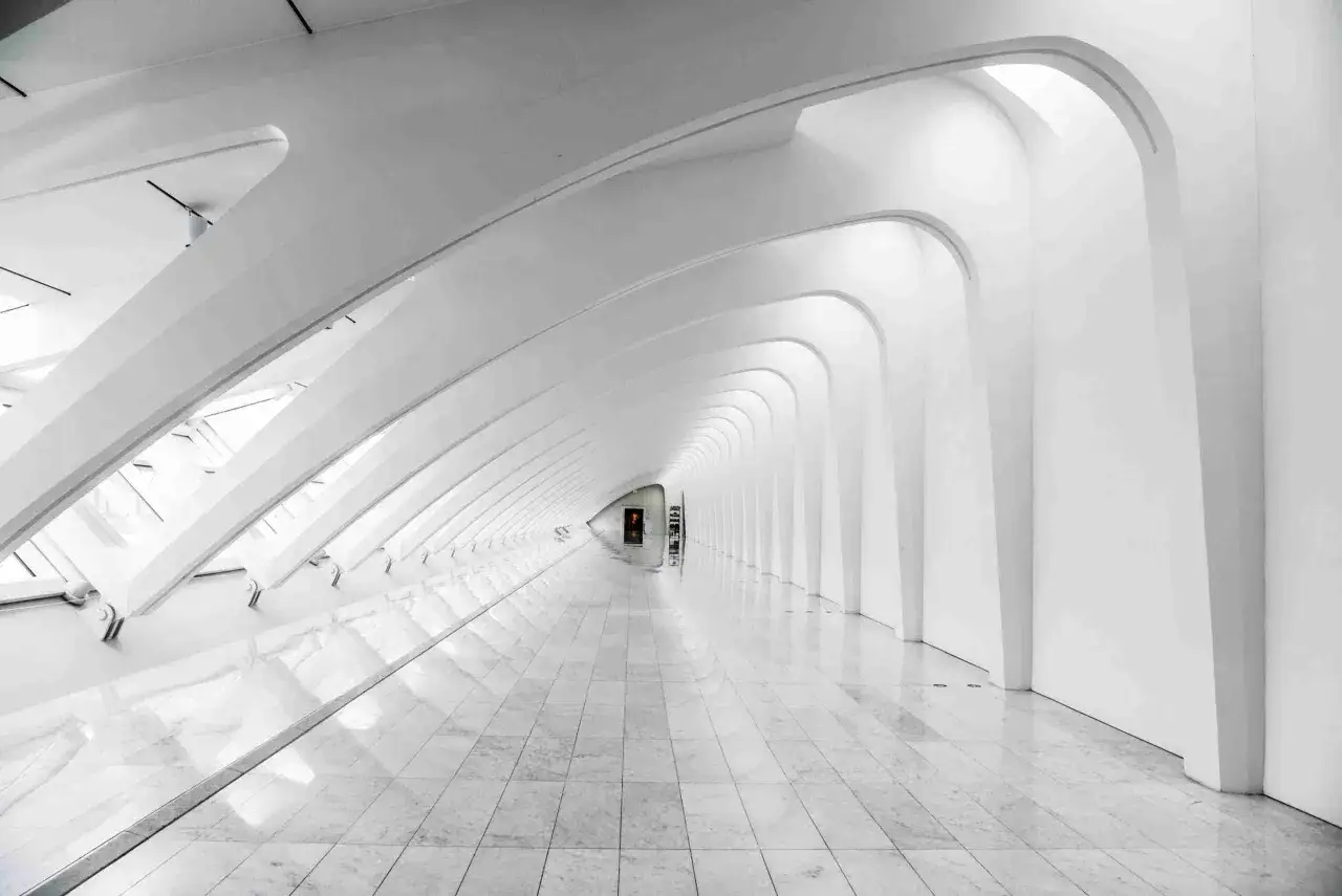 Corridoio bianco e luminoso con archi ripetuti, pavimenti in marmo lucido. La **less is more traduzione** si percepisce nell'essenzialit&agrave; delle forme.
