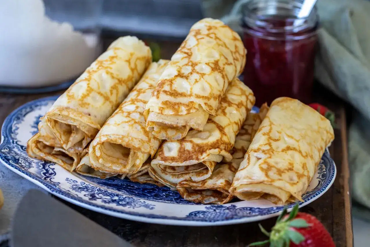 En hög med gyllene, glutenfria pannkakor rullade och staplade på en blåvit tallrik, med sylt och jordgubbar i bakgrunden.
