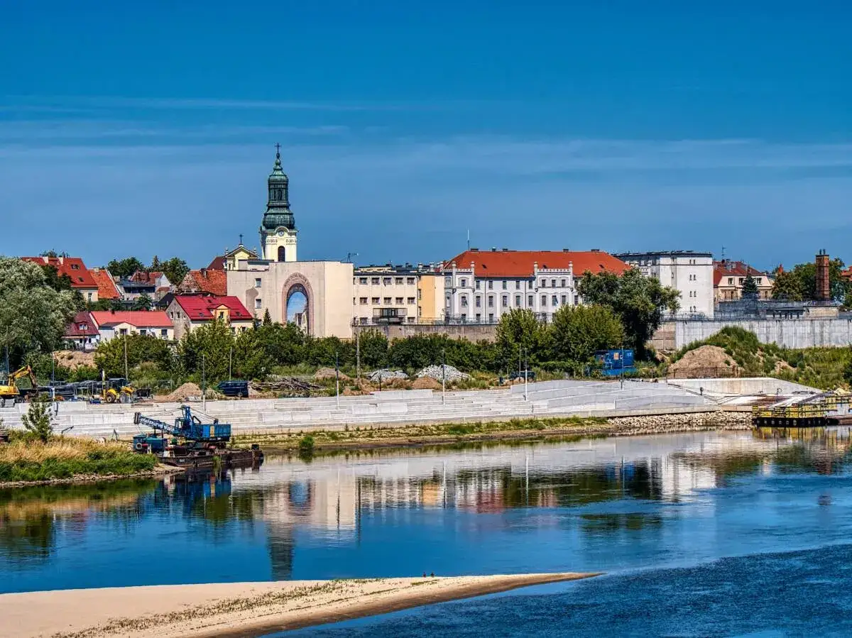 Widok na miasto z wieżą kościelną i rzeką w tle. Widać budynki z czerwonymi dachami, plac budowy z dźwigiem i piasek. Fordon w słońcu.