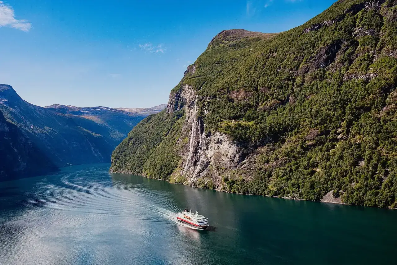 Traumhaftes Wetter in Norwegen im August: Ein Kreuzfahrtschiff gleitet durch einen Fjord, umgeben von steilen, bewaldeten Bergh&auml;ngen und einem Wasserfall.