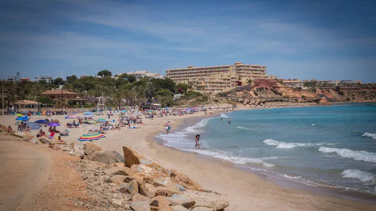 Gente disfrutando del sol y el mar en la animada campoamor playa, con edificios tur&iacute;sticos al fondo.