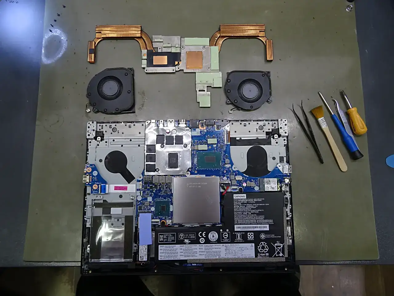 Rozłożone części laptopa: płyta gł&oacute;wna, wentylatory, radiator, bateria i narzędzia do naprawy.