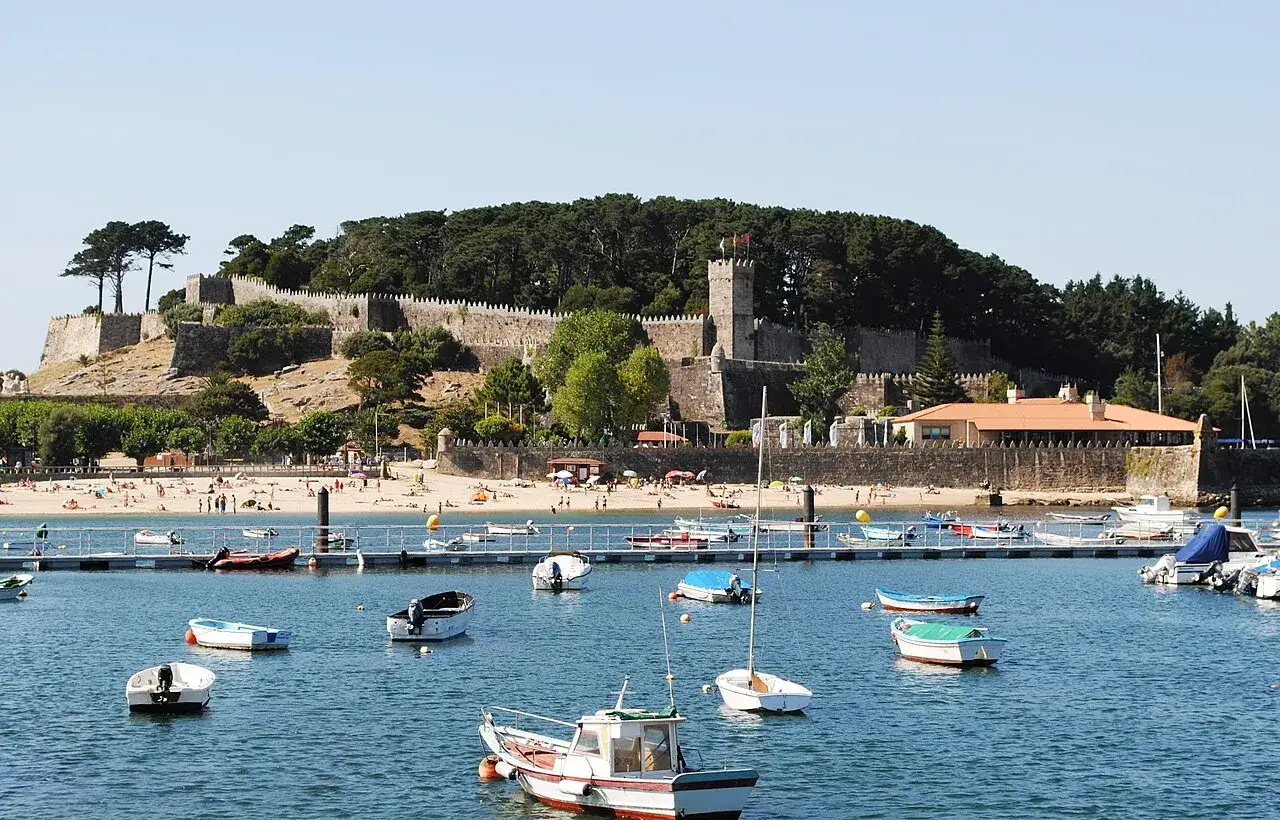La **fortaleza de Monterreal** se alza majestuosa sobre una colina, vigilando un puerto lleno de barcas y una playa con gente disfrutando del sol.