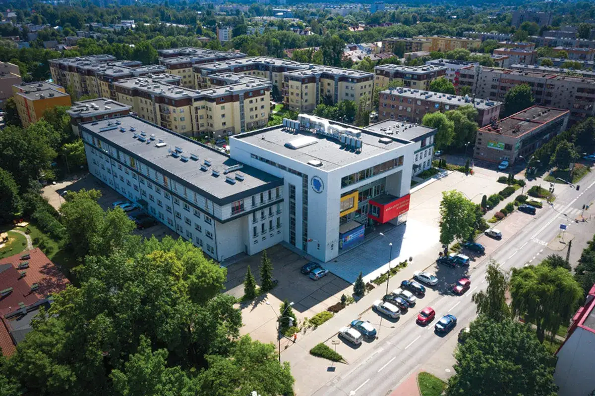 Nowoczesny budynek Akademii Śląskiej otoczony zielenią, z parkingiem i ulicą w tle.