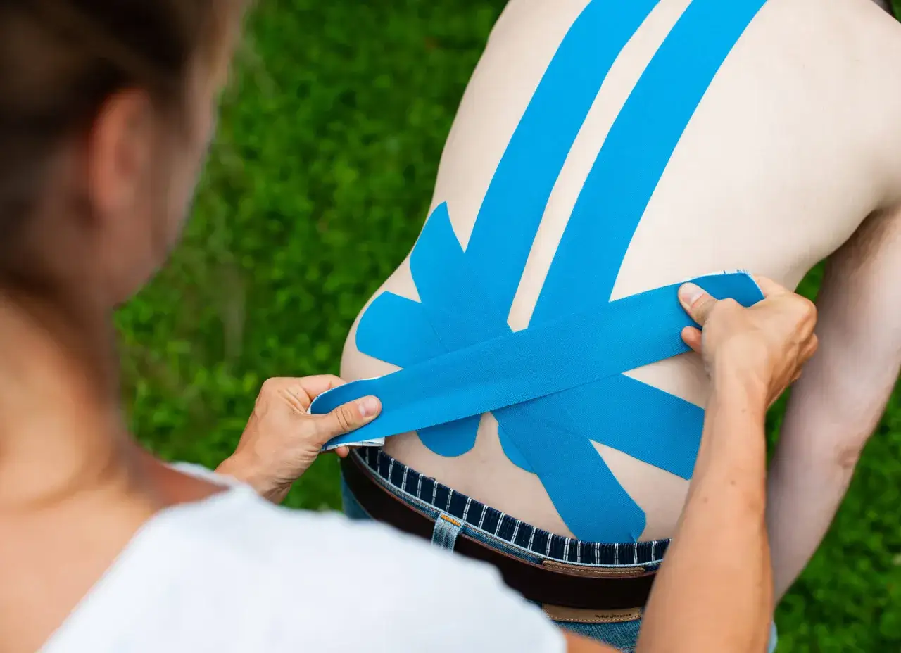 Eine Person legt blaues Kinesio-Tape auf den R&uuml;cken eines Mannes, um LWS-Beschwerden zu lindern.