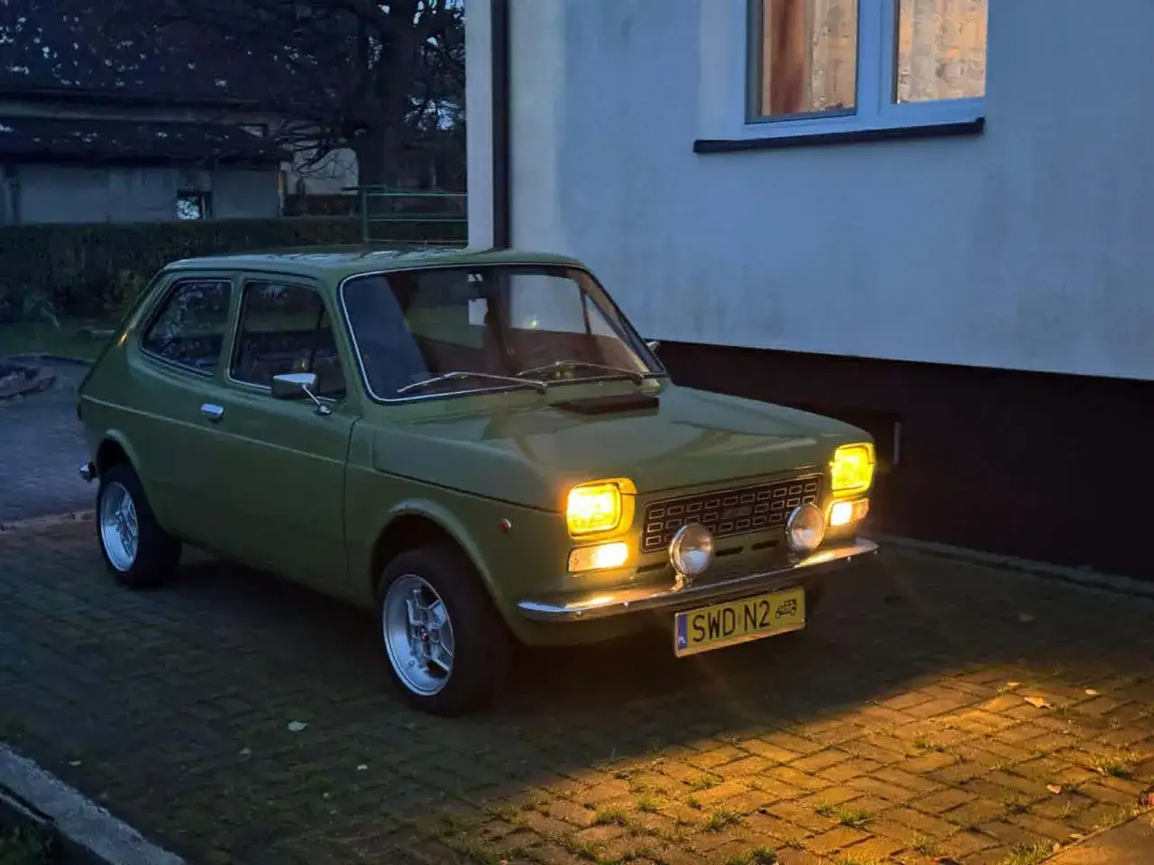 Zielony Fiat 127 z lat 70. stoi na podjeździe, jego światła rozświetlają zmierzch.