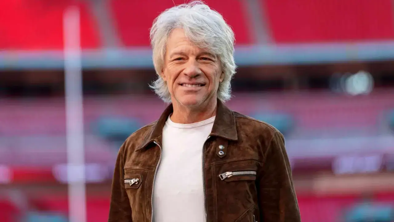 Jon Bon Jovi, w brązowej kurtce, uśmiecha się przed stadionem, gotowy na kolejny bon jovi koncert.