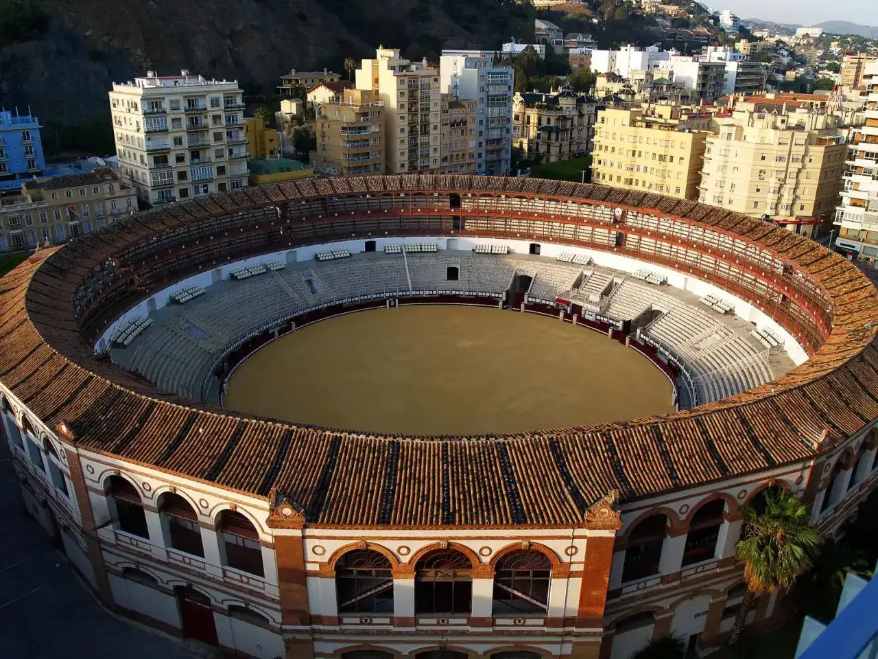 Vista a&eacute;rea de la plaza de toros de M&aacute;laga, con su arena circular y gradas, rodeada de edificios urbanos.