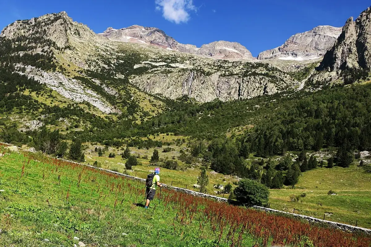 Senderista camina por un prado verde hacia imponentes montañas. A lo lejos, se vislumbra una cascada. El paisaje evoca la paz de una cabaña de turmo.