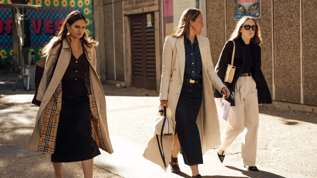 Tres mujeres pasean con estilo urbano, una con gabardina de cuadros, otra con blazer crema y falda vaquera, y la tercera con chaqueta negra. Un toque gorpcore moderno.