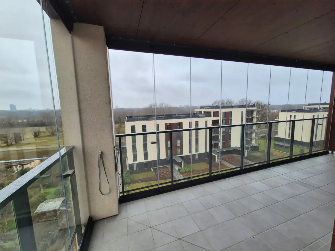 Zabudowa balkonu w bloku z widokiem na nowoczesne osiedle. Szklane panele tworzą przestronną, przeszkloną loggię.