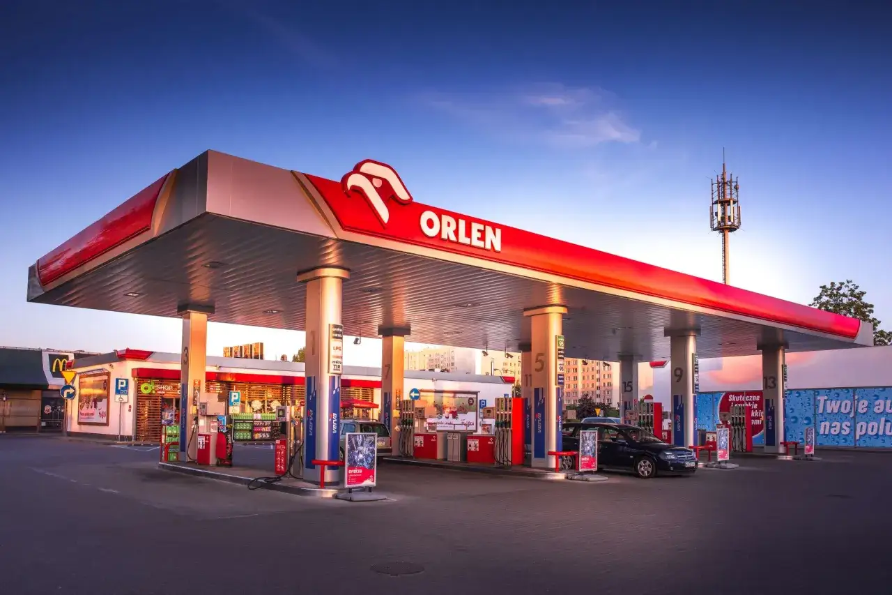 Stacja Orlen z logo i czerwonym zadaszeniem. Samochody tankują paliwo, a obok znajduje się sklep. Idealne miejsce na orlen promocja na paliwo.