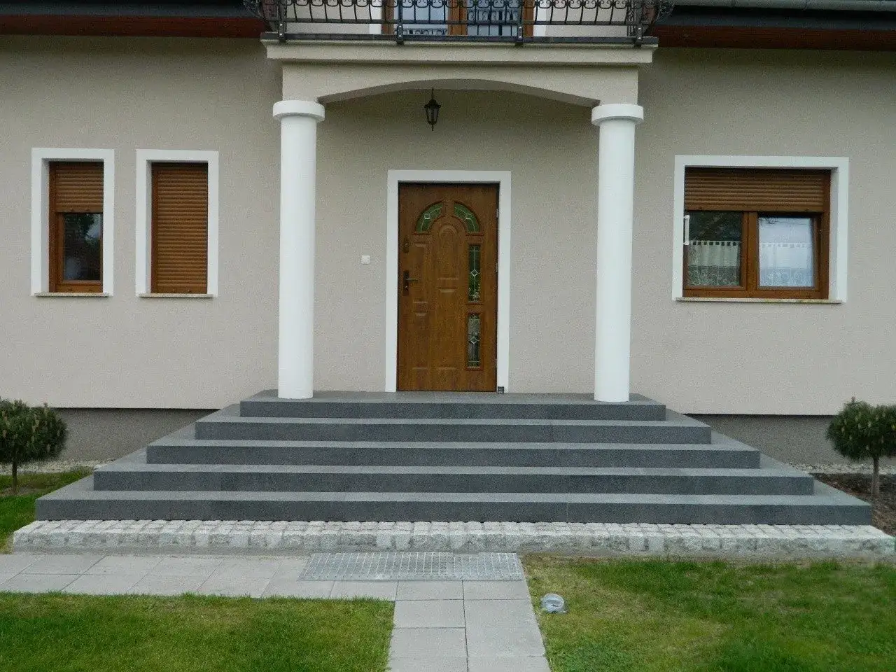 Eleganckie, wygodne schody prowadzą do drzwi wejściowych. Dwa białe filary podtrzymują daszek.