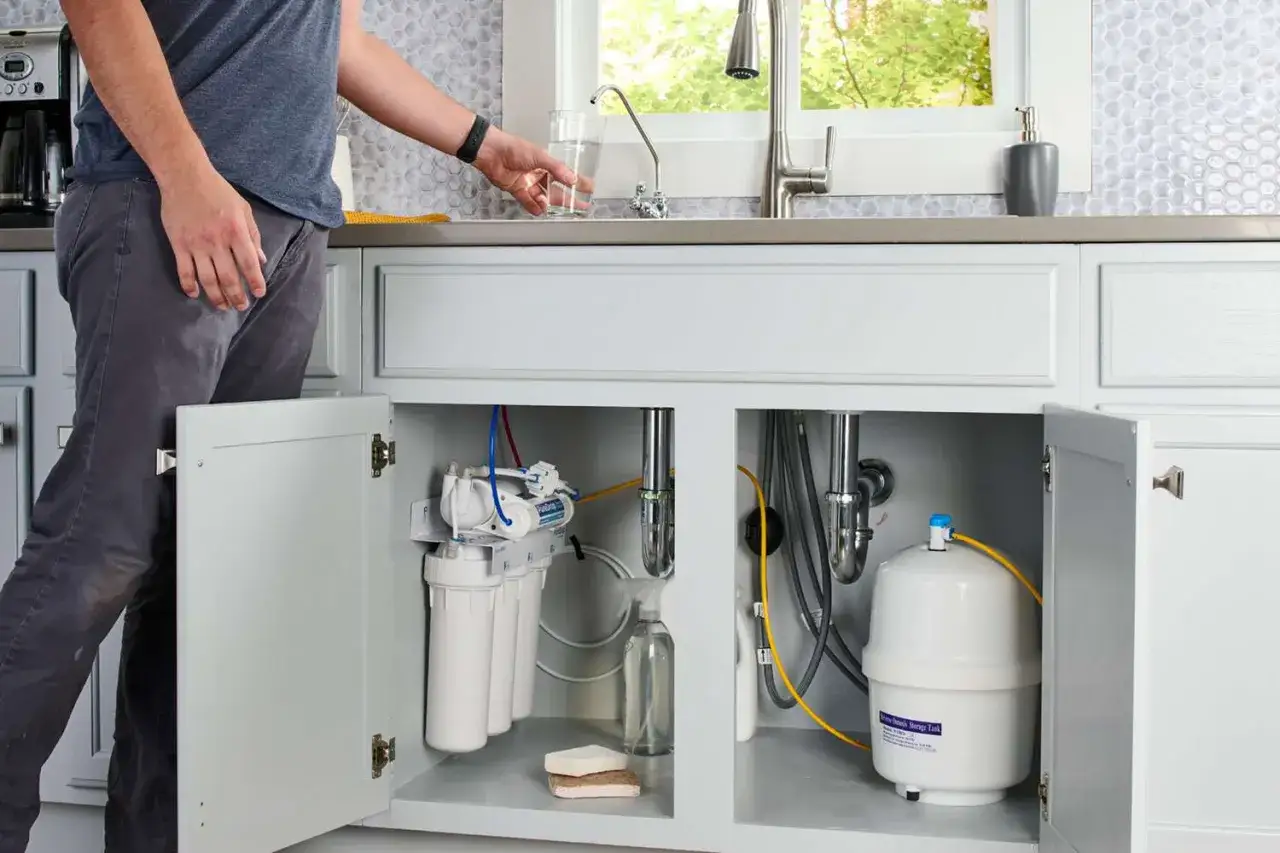 Sous l'évier, un système de filtration d'eau osmosée est installé. Un homme se sert un verre d'eau pure.