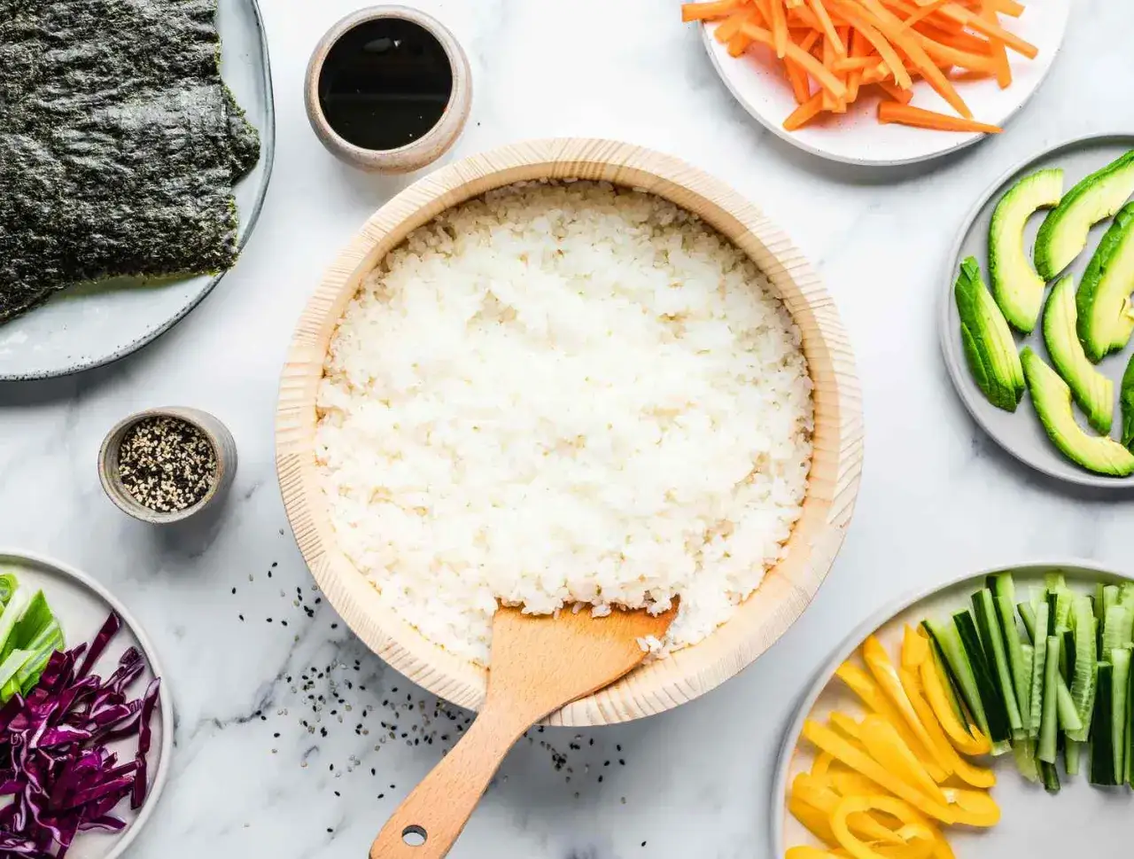 Gotowy ryż do sushi w drewnianej misce, obok nori, awokado, marchewka, ogórek, papryka, cebulka i sos sojowy.