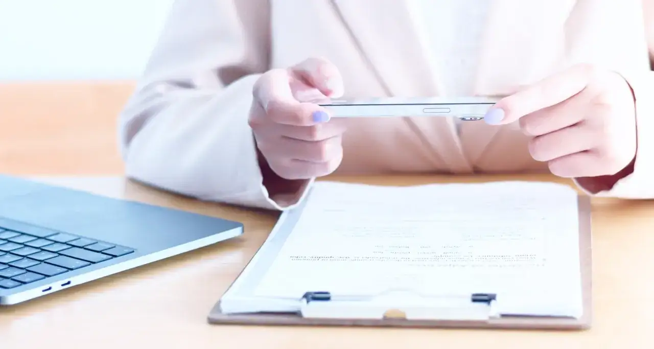 Dłoń trzyma telefon, używając go jako skaner w telefonie do dokumentu na desce. Obok leży laptop.