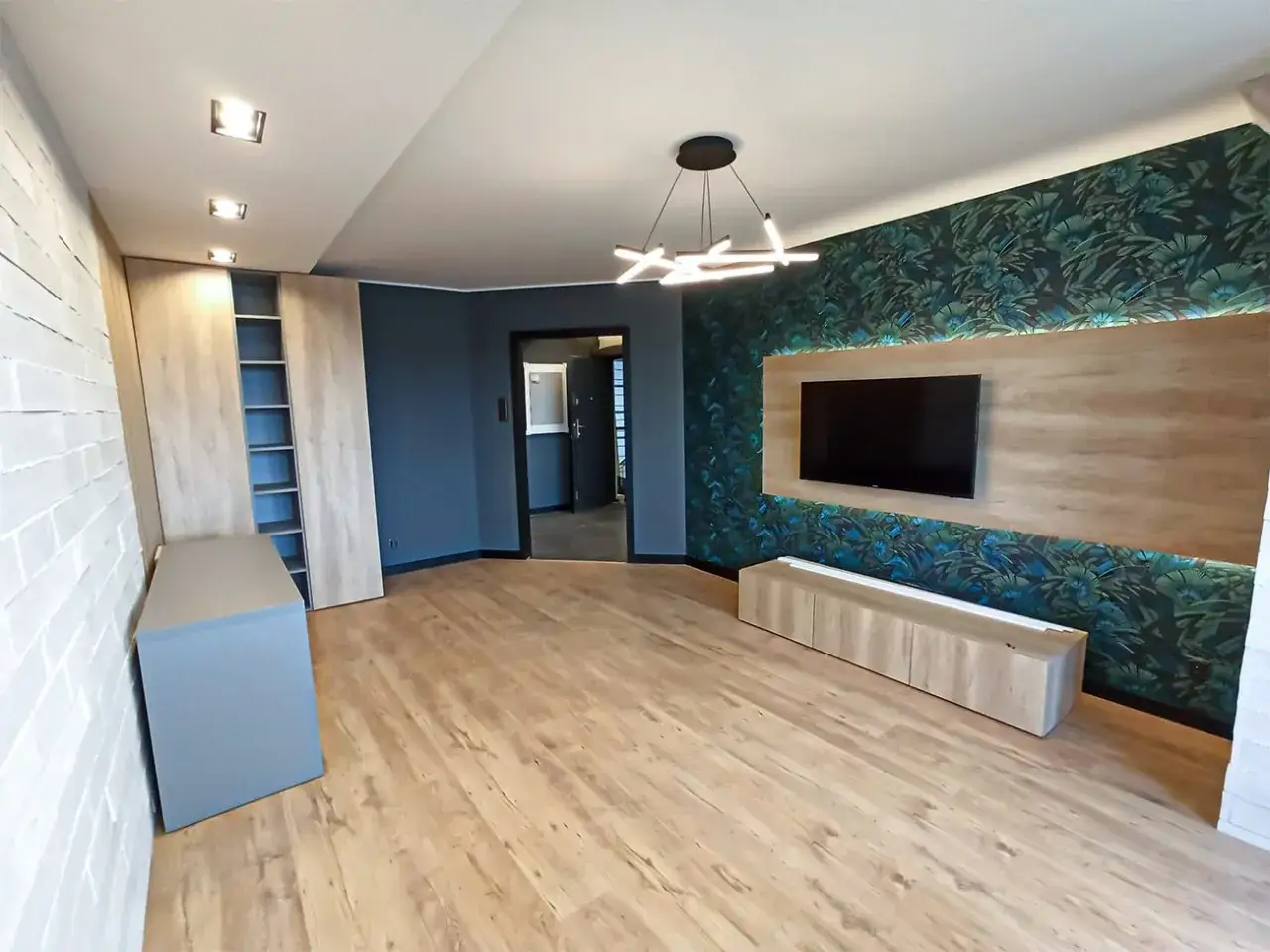 Nowoczesny salon z telewizorem na tle tapety z motywem liści, drewnianą zabudową i oświetleniem.
