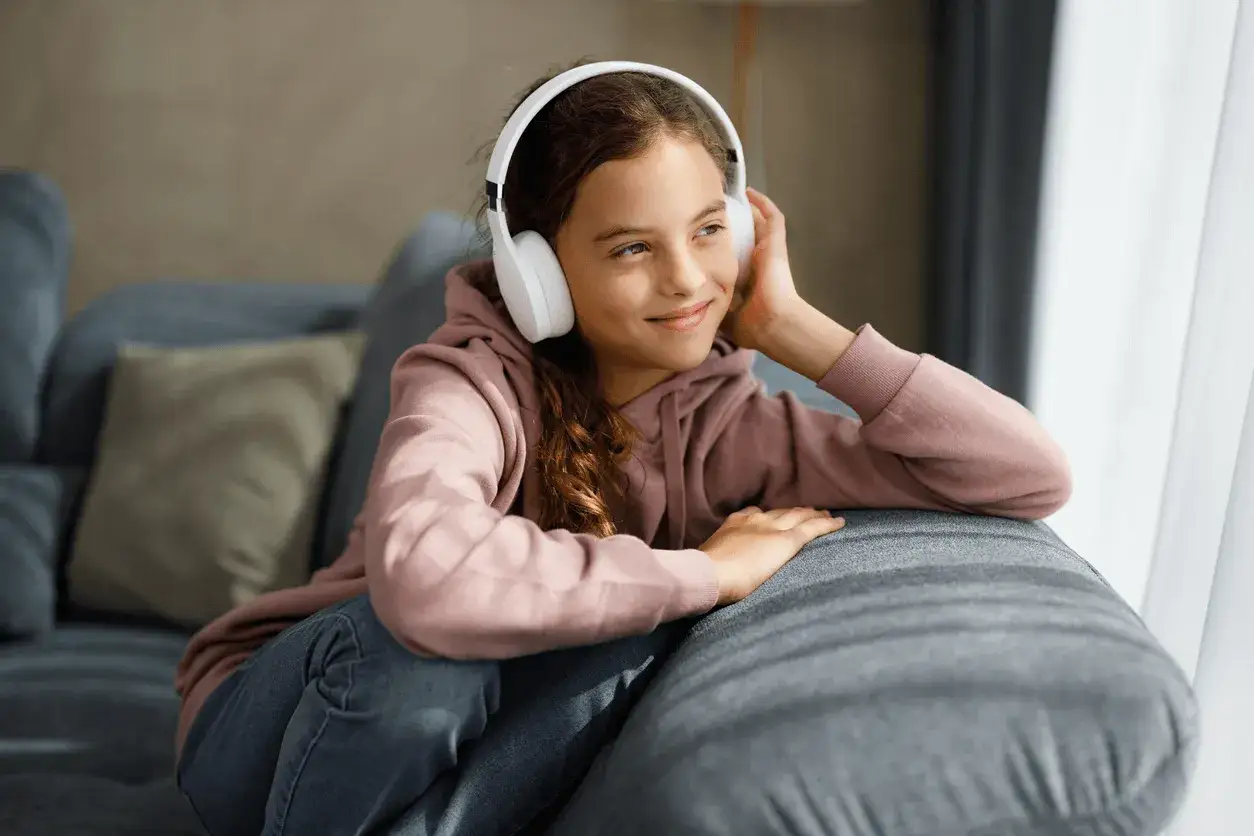 Dziewczynka w słuchawkach słucha audiobook&oacute;w po angielsku z tekstem, relaksując się na kanapie.