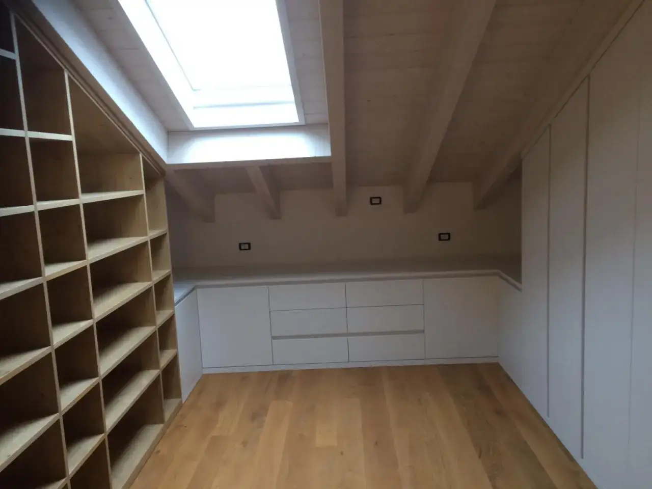 Ampia libreria in mansarda con lucernario e mobili contenitori bianchi.