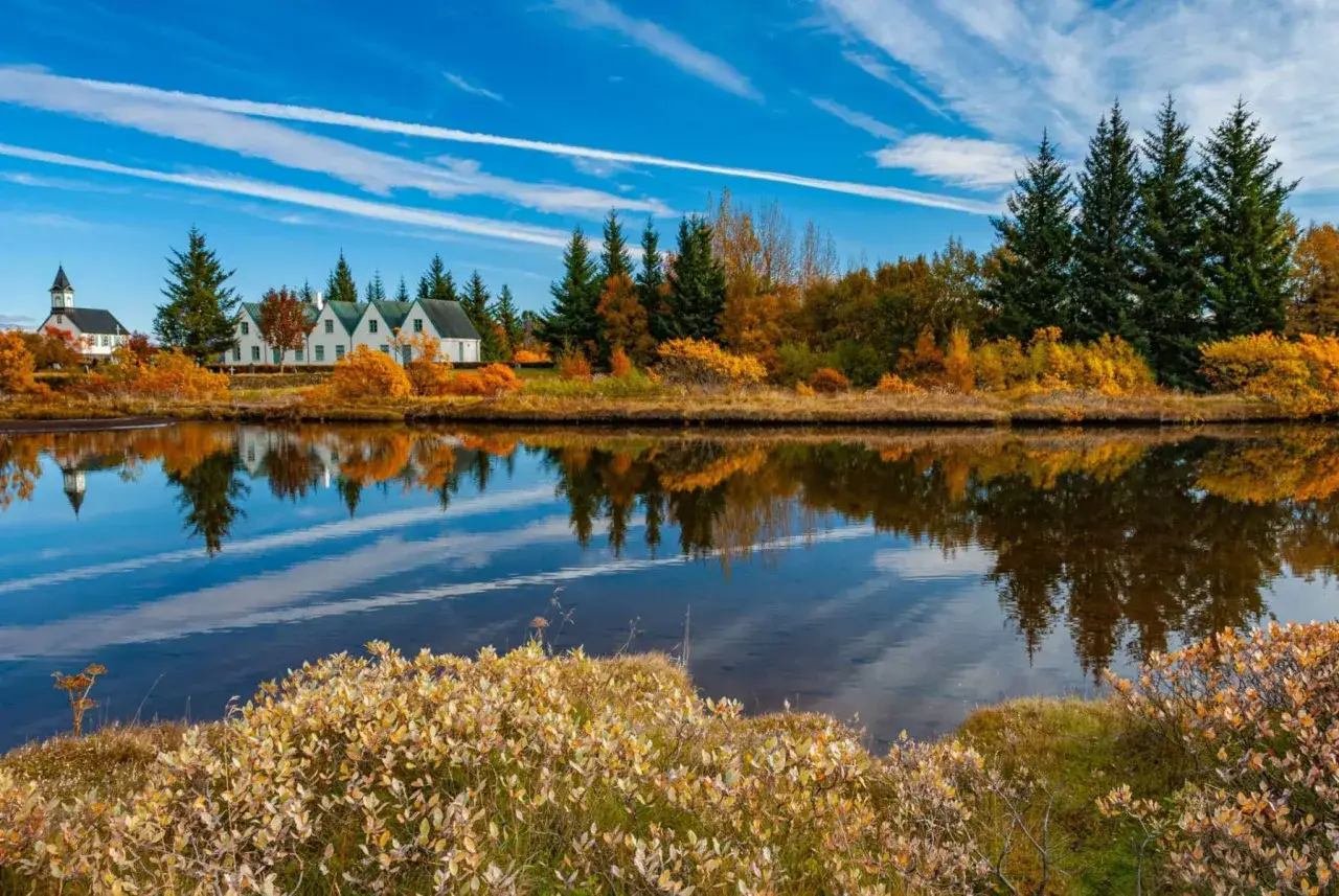 Herbstliches Wetter auf Island im September: bunte Bäume spiegeln sich im ruhigen Wasser, Häuser und Kirche am Ufer.