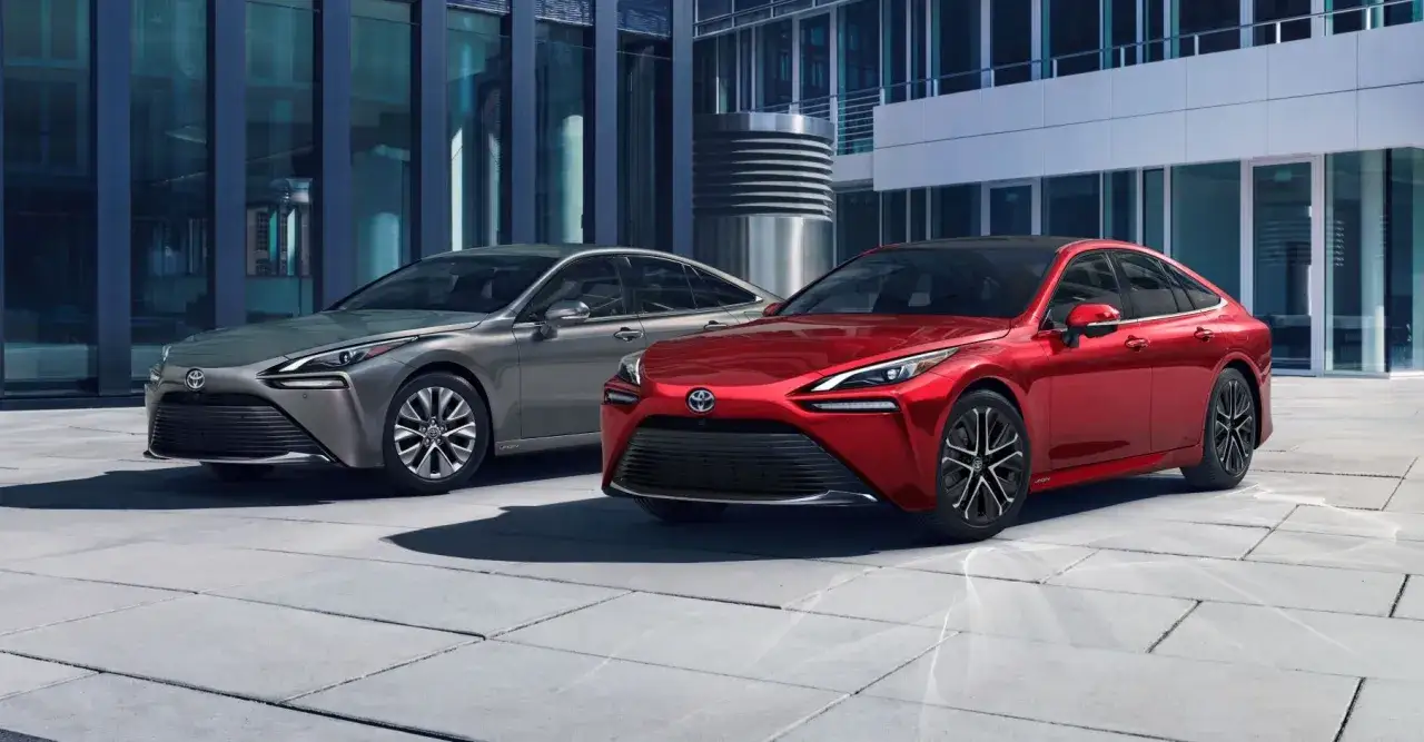 Dwa samochody Toyota Mirai, jeden czerwony, drugi srebrny, stoją przed nowoczesnym budynkiem. Sprawdź, jaka jest toyota mirai cena.