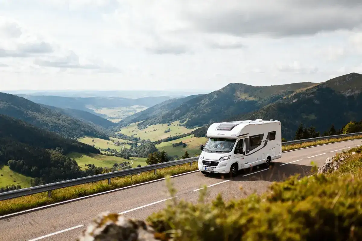 Un camping-car blanc sillonne une route de montagne sinueuse, offrant des vues spectaculaires sur des vallées verdoyantes. L'un des plus beaux circuits camping-car.