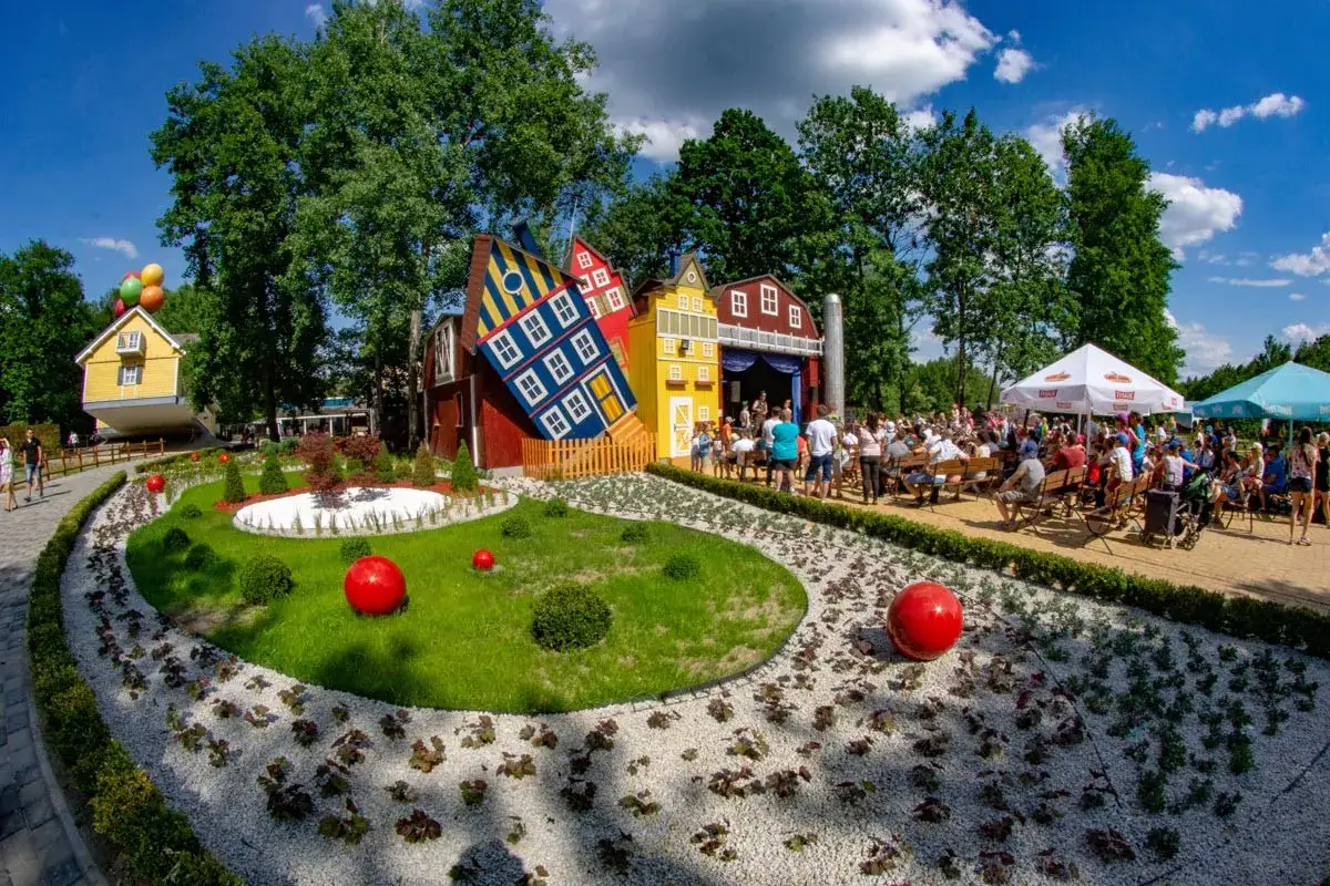 Kolorowy park rozrywki z krzywymi domkami i sceną, oferujący atrakcje dla dzieci zachodniopomorskie. Tłum widzów siedzi na ławkach.