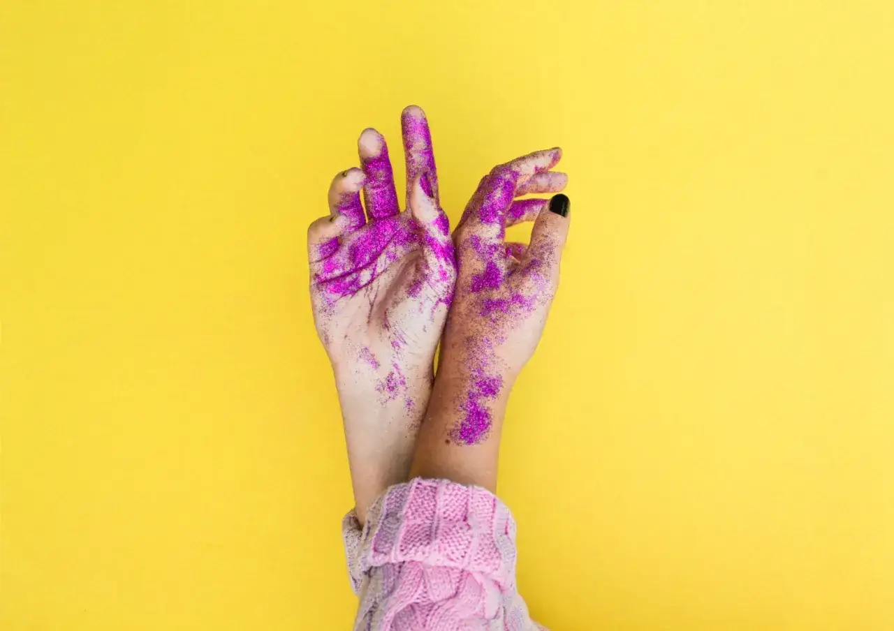 Mani cosparse di glitter viola su sfondo giallo, il colore complementare del giallo, che creano un contrasto vibrante.
