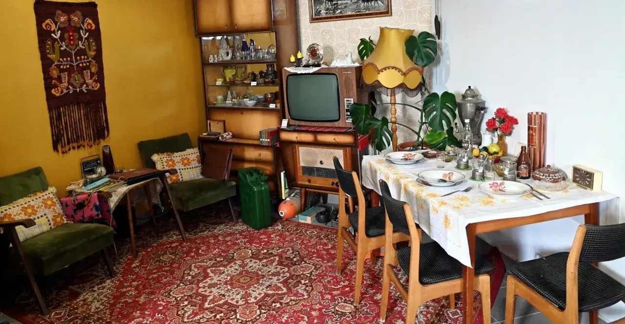 Salon w stylu prl: zielone fotele, dywan, meblościanka, telewizor, radio, stół z zastawą i kwiatami.