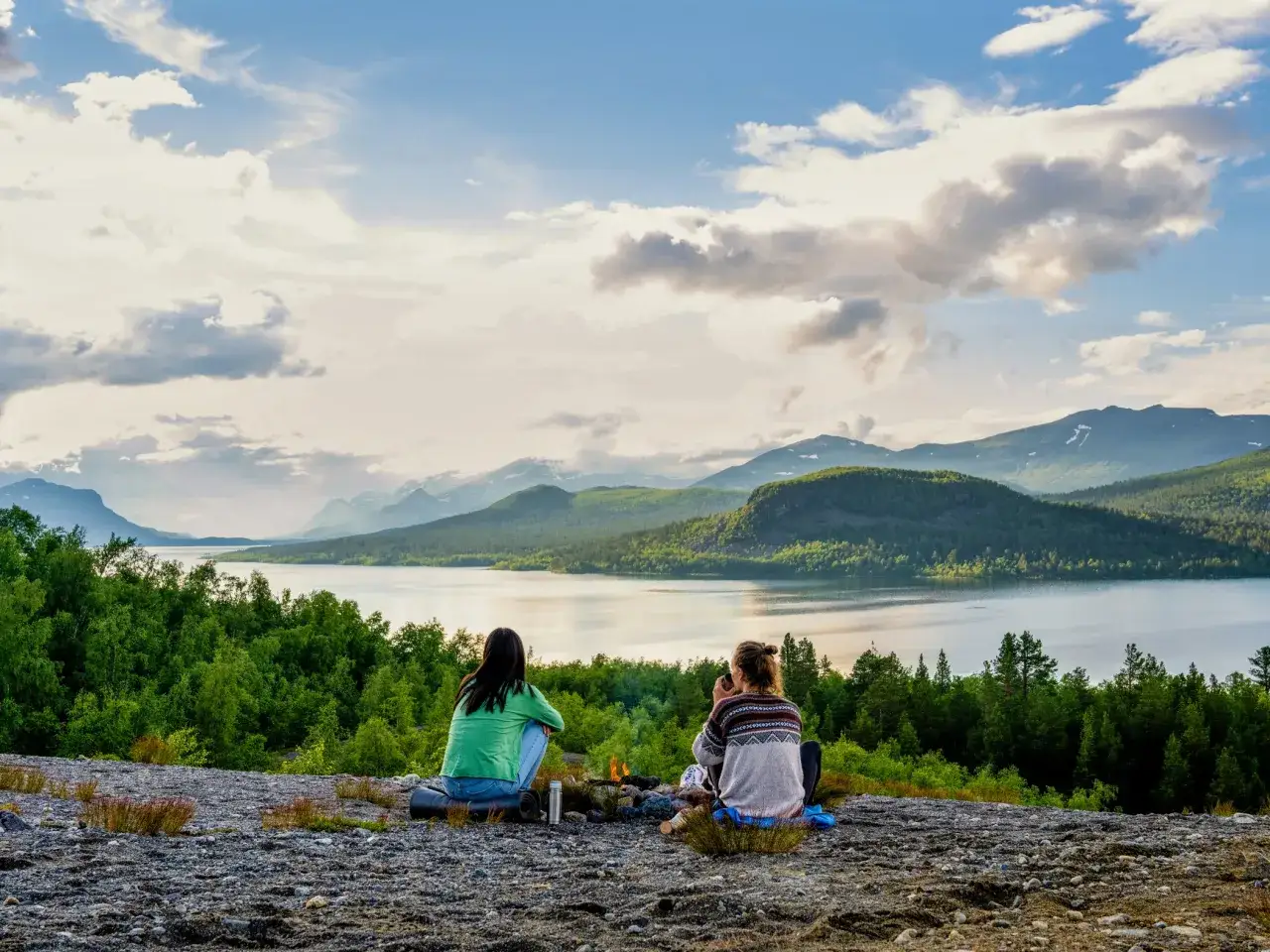 Tv&aring; v&auml;nner sitter vid en eld brasa och njuter av utsikten &ouml;ver en sj&ouml; och berg.