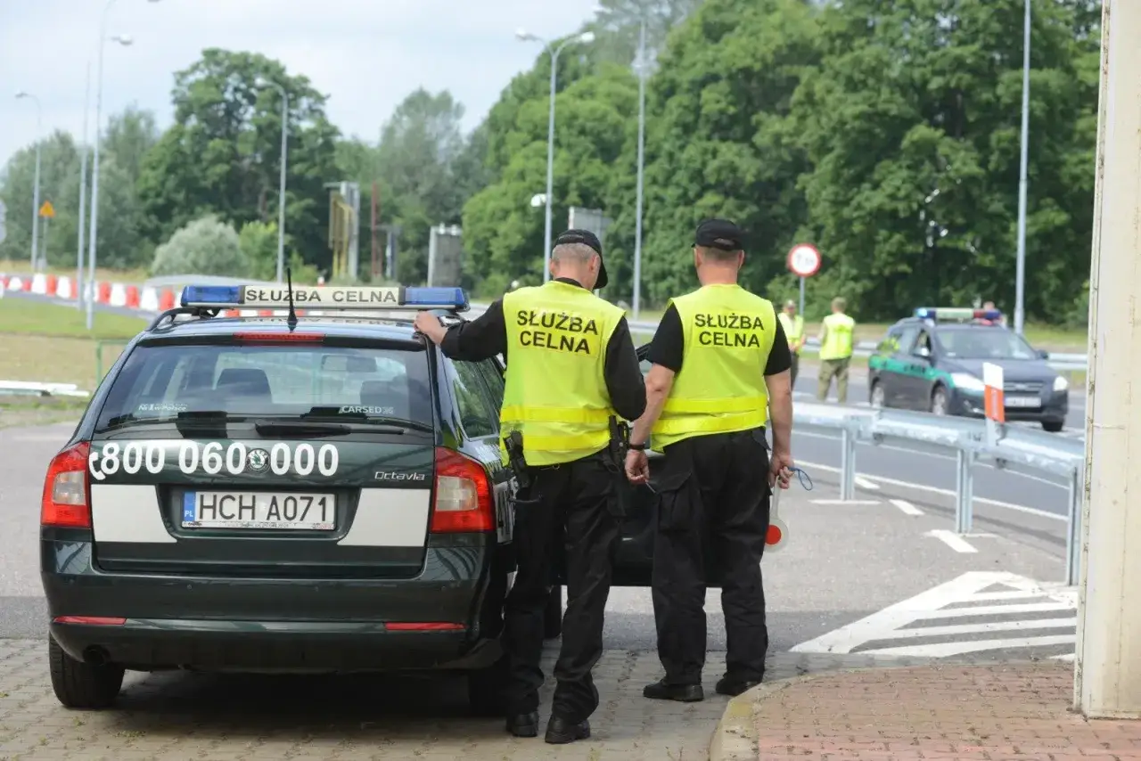 Służba celno skarbowa kontroluje ruch na drodze. Dwóch funkcjonariuszy w kamizelkach odblaskowych stoi przy oznakowanym radiowozie.