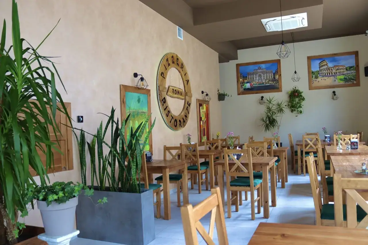 Wnętrze restauracji Pizzeria Roma. Drewniane stoły i krzesła, ozdoby ścienne z motywami Rzymu, dużo zieleni.